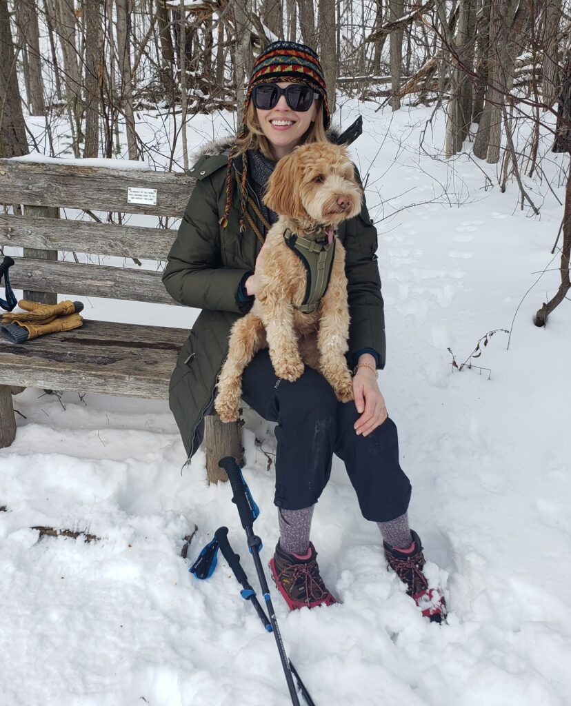 Gillian Gillies seated on a snowy trail with her puppy Reba during a winter hike